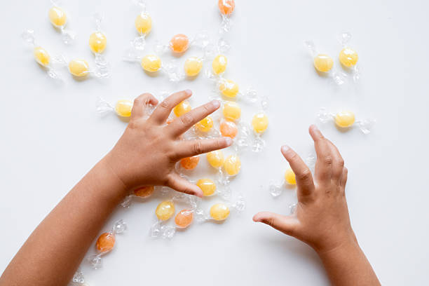Child hands reaching for wrapped yellow and orange hard candies on white surface. High quality.