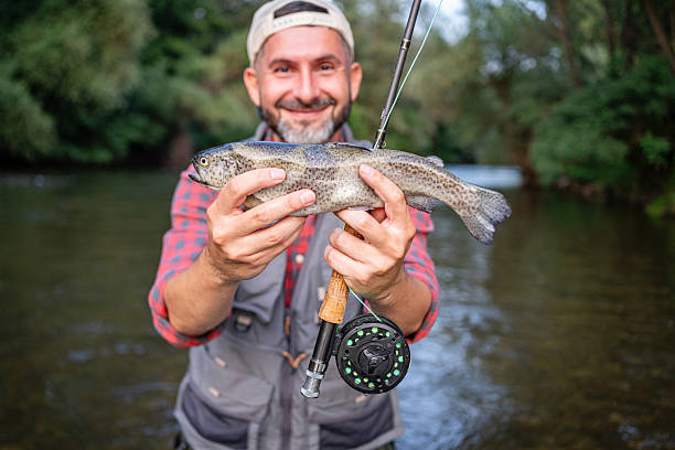 A smiling man proudly displays a freshly caught trout while fly fishing in a beautiful, forested river landscape, showcasing an exciting outdoor adventure.