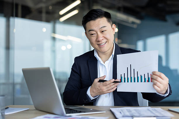 istock Asian young man, office worker holding a video conference, talking and showing documents with graphs to the camera 2226122827