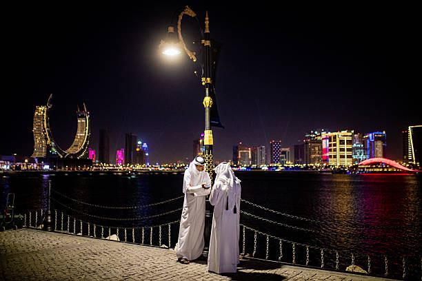 Two Arab men stand under streetlights by Lusail's shoreline, engaged in conversation, Qatar.
