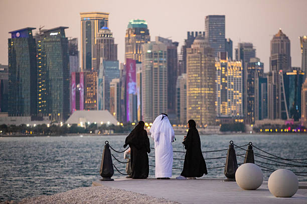 An Arab man and two  Arab women stand on the beach, gazing at the skyscrapers across the river, Qatar.