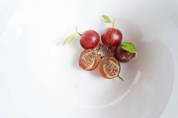 several ripe gooseberries arranged in a white bowl. One berry is cut in half, revealing its flesh and seeds. The background is a clean, white bowl