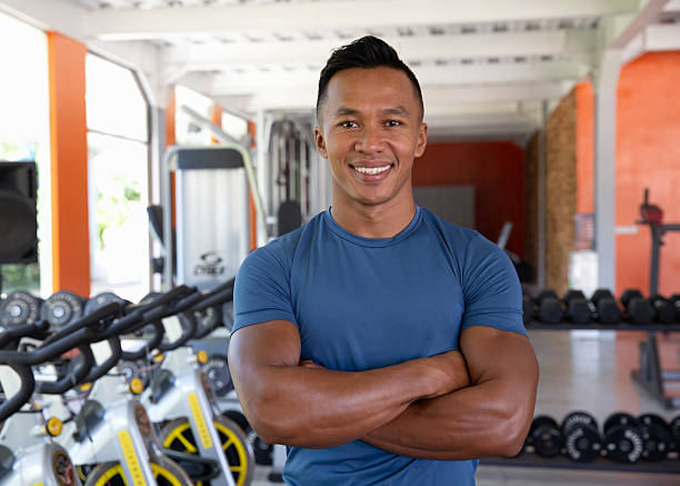 Close-up shot of a smiling male fitness trainer standing confidently with arms crossed inside a modern gym, surrounded by workout equipment and exercise bikes.\n\nConcept:\nconfident personal trainer, fitness coach, gym workout, bodybuilding, athletic man, fit male