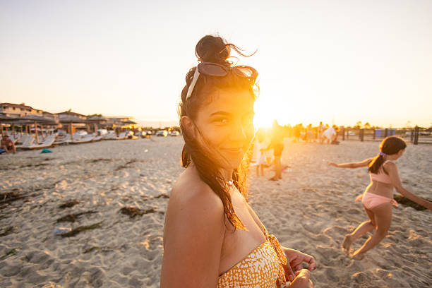 A young woman with a radiant smile looks towards the camera, her hair gently blowing in the wind, as the golden sun sets behind her on a sandy beach.