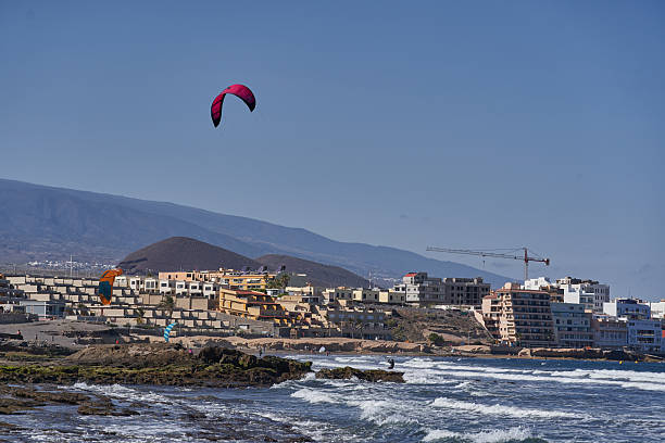 Spain, Canary Islands, Tenerife, Playa del Médano 05 June 2025 - Kitesurfers riding near rocky coastline with colorful canopies and urban skyline in background