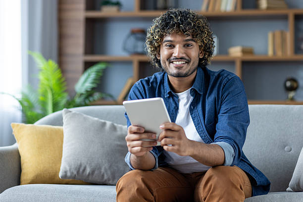 istock A happy man with curly hair sits on a couch, using a tablet indoors at home. 2226143435