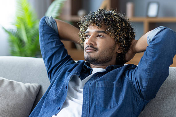 istock A relaxed young man sits on a couch with hands behind his head, deep in thought. 2226145587