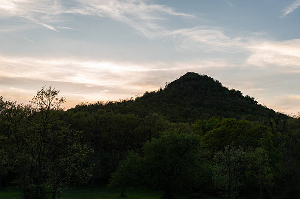 A conical volcanic Hegyesd Hill blanketed with dense forest is silhouetted against a colorful sunset sky. A grassy clearing in the foreground adds depth to the tranquil natural scene, while a contrail streaks across the evening clouds.