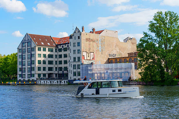 Berlin, Germany - May 30, 2025: Modern boat travels along the Spree River in Berlin, showcasing historic buildings with unique graffiti under a bright blue sky during a sunny day.