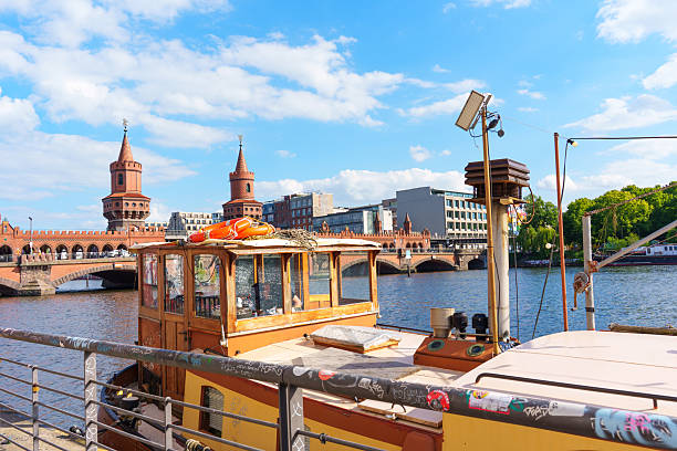 Berlin, Germany - May 30, 2025: Wide-angle view of the Oberbaum Bridge in Berlin, featuring a boat in the foreground and the city skyline under a bright blue sky with fluffy clouds.
