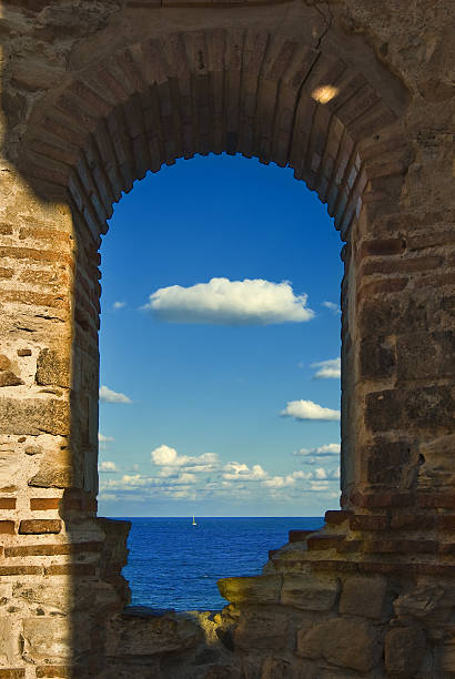 Sail boat, calm sea, blue sky and white clouds seen through the stone window of the old castle in summer.
