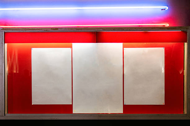 Three blank white posters pinned on a red display board, illuminated by colorful neon lights in blue and pink tones.