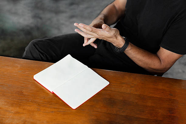 A man in black outfit sitting at a wooden table with an open blank notebook. His hands are mid-gesture, suggesting thought, explanation, or decision-making. A conceptual image about planning, brainstorming, or communication, with space for text or design overlay.