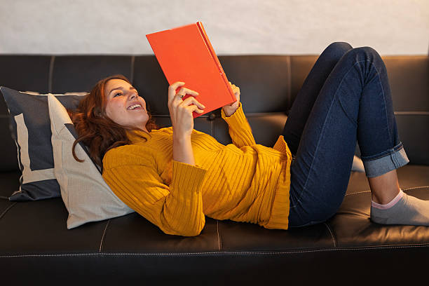 istock Young woman lying on sofa reading a book and smiling 2226237222
