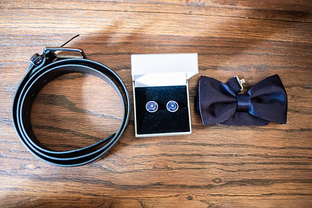 A stylish collection of wedding accessories features a black belt, elegant cufflinks in a box, and a sleek bow tie, perfect for the groom.