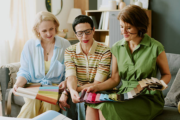 Three middle aged Caucasian women sitting together examining fabric swatches, discussing textile samples and collaborating on design choices in modern living room setting