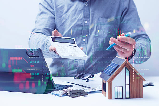 istock Businessman holding calculator with house model on table. Tablet with dollar bills with digital overlay of stock market charts and uptrend line. 2226242780