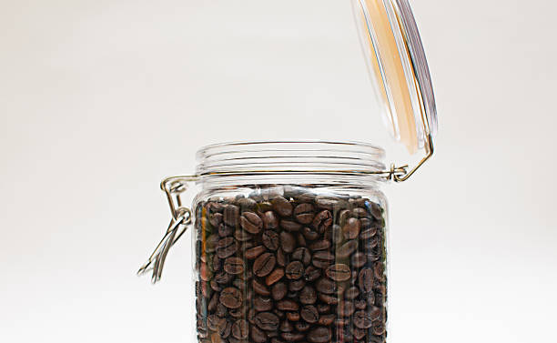 Coffee beans packed in a clear glass jar with a lid open close-up white background