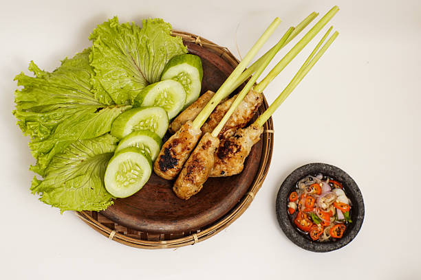 Traditional Balinese satay lilit served on lemongrass sticks with fresh lettuce, cucumber slices, and spicy sambal matah in a stone mortar. Isolated on white background.