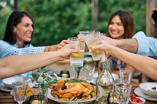 A group of friends toasting with drinks and enjoying a homemade meal outdoors. The cheerful atmosphere features fresh food, greenery, and shared laughter.