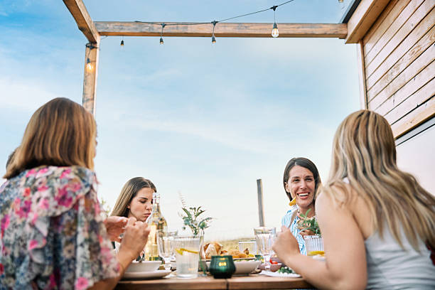 A group of women enjoying a relaxed outdoor lunch with homemade dishes, set in nature under a clear sky, near a rustic cottage.