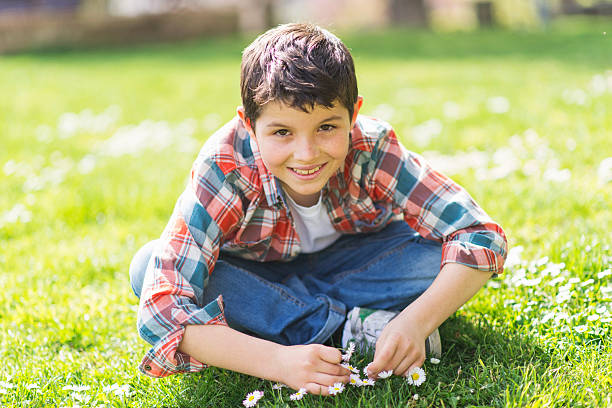 A young Caucasian boy with short dark hair, wearing a plaid shirt, jeans, and sneakers, sits cross-legged on grass in a sunlit park, smiling cheerfully.