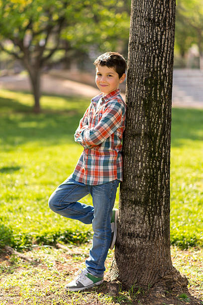 istock Smiling boy in plaid shirt leaning against a tree in a park. 2226248293
