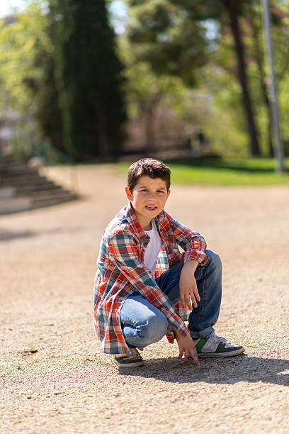 A young Caucasian boy with short dark hair, wearing a plaid shirt, jeans, and sneakers, squats on a gravel path in a sunlit park, looking curious.