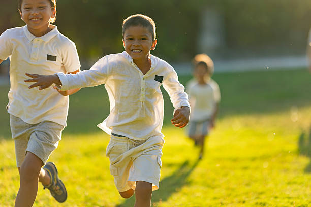 A small group of young children are seen running down a grassy hill with their arms out like airplanes as they enjoy the fresh air.  They are each dressed casually and are laughing and smiling.