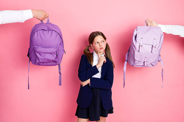 istock Charming schoolgirl in uniform making a choice between two backpacks on a bright pink background 2226278547