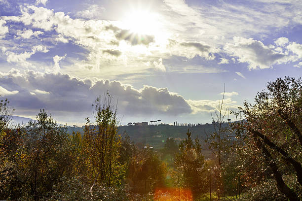 Tuscany, Italy – Autumn scenic view of Tuscan countryside with silhouettes of cypress trees and villas on a hill illuminated by bright rays of sun breaking through the clouds. Typical Italian rural landscape with natural colors and sun reflections.