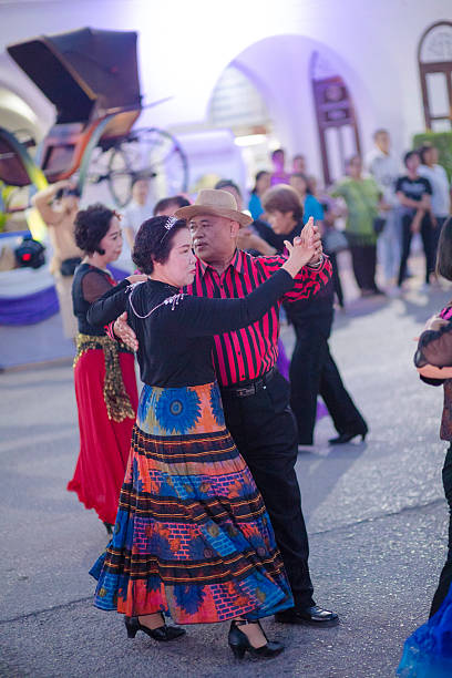 Group of dancing mature thai couples. People are  captured at night market in public. People are dancing among other mature thai people who are dancing classic spanish dances. In background are visitors of night market. People are wearing classic dance fashion. Woman is wearing a colorful skirt and man a red and black shirt and a hat . Scene is at night market in Lampang. People are dancing in public to public music by band. Market is near train station on small square. Lampang is city in northern Thailand south of Chiang Mai