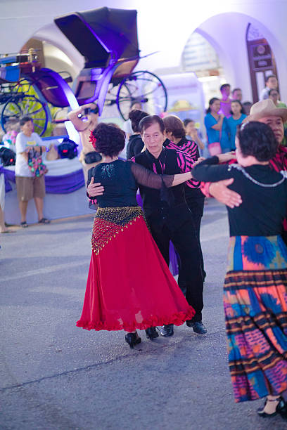 Dancing mature thai couples in front of old carriage. People are  captured at night market in public. People are dancing among other mature thai people who are dancing classic spanish dances. In background is a building and an old carriage is placed on a podest. People are wearing classic dance fashion.  Scene is at night market in Lampang. People are dancing in public to public music by band. Market is near train station on small square. Lampang is city in northern Thailand south of Chiang Mai and is also city for horse carriage as public tour transportation