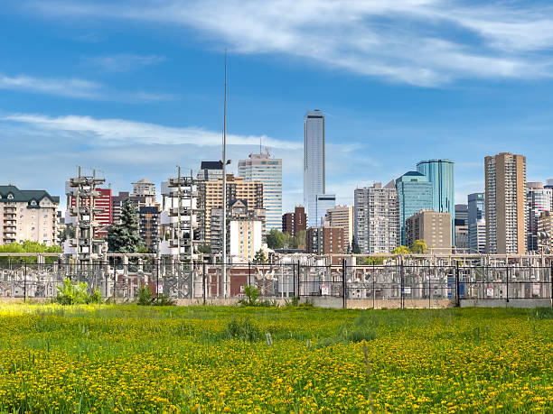 Commercial image showing Edmonton’s downtown high-rise skyline framed by a high-voltage electrical substation in the foreground. The fenced transformer yard sits against a lush green field under a bright blue sky, illustrating the intersection of urban growth and energy infrastructure in Alberta’s capital city.