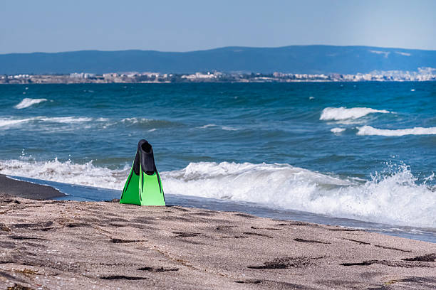 Bright green swimming fins sit on the soft sand at a warm summer beach, while waves roll in from the sea. Background. Sport, activity, leisure.