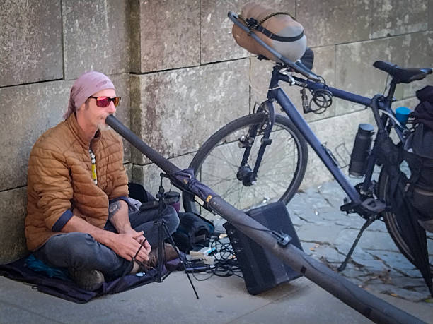 Porto, Portugal: August 12 2023: Front view, medium shot of a street musician sitting on the ground playing a didgeridoo next to two parked bicycles.