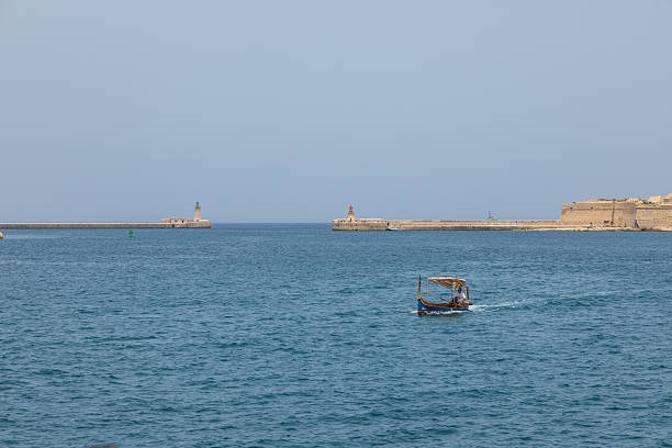 Valletta, Malta – June 21, 2021: A man on a traditional boat with a red canopy in the harbor, historic buildings and yachts in the background.