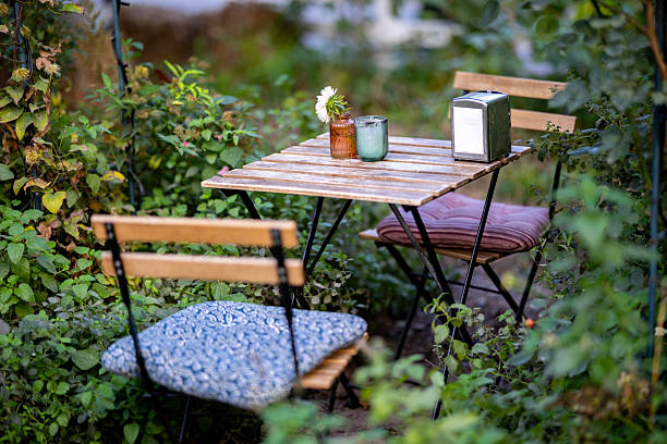istock A cozy cafe setting with a small wooden table and two chairs featuring cushions, nestled amongst lush greenery, with a single white flower in a vase, a green glass, and a napkin holder on the table. 2226349223