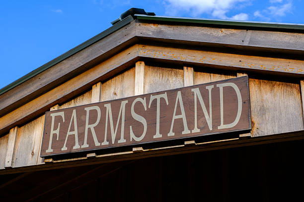 A wooden sign reading FARMSTAND mounted on the gable of a rustic wooden building, set against a blue sky. The image evokes rural charm and seasonal agriculture.