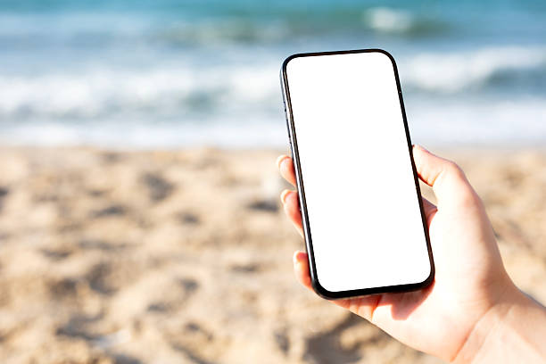 A woman holding black mobile phone with blank desktop screen while sitting on a beach