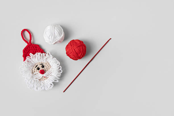 Crocheted toy Santa Claus in a red hat with balls of thread and crochet hook on gray background. Baby stuff and accessories. Gift for winter holidays. Top view, flat lay.