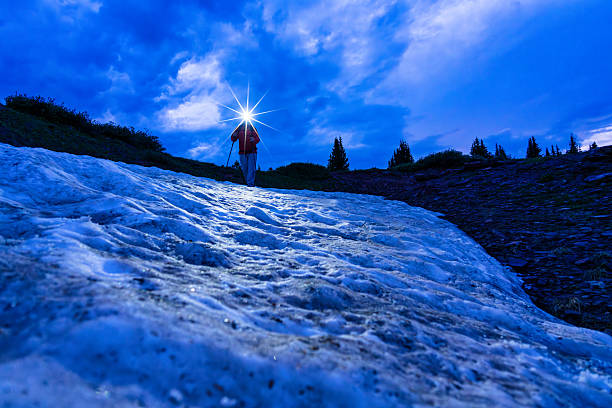 A lone hiker descends a snowfield at dusk under a dramatic blue sky, illuminated by a bright headlamp. The scene conveys themes of adventure, solitude, determination, and exploration in a remote alpine environment. Shot in low light with a wide-angle perspective and starburst effect from the headlamp for added drama and atmosphere.. Vail, Colorado USA.