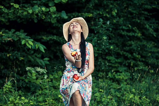A cute girl with two red apples in the forest. A woman in a floral dress and red apples. A seductive girl against the backdrop of green trees.