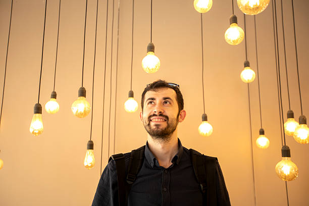 A smiling man with a beard stands under many hanging light bulbs, looking up. The warm lights create a cozy and creative atmosphere. He wears a dark shirt and a backpack.