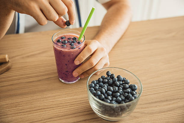 Young man preparing blueberry smoothie on wooden table in kitchen at home.