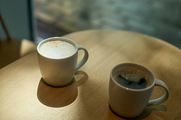 Two mugs filled with different types of coffee sit on a wooden table, illuminated by sunlight streaming through a nearby window