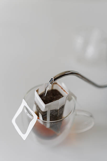 Boiling water pouring from kettle into drip coffee bag placed in glass cup, offering simple and convenient way to brew fresh cup of coffee, close-up.