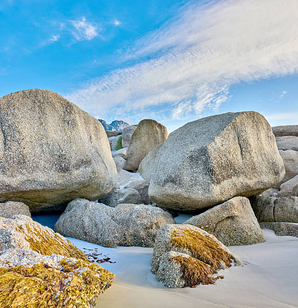 Large granite boulders resting on a sandy beach create a stunning natural landscape. The bright blue sky adorned with scattered clouds adds a serene and picturesque backdrop to this coastal scene.
