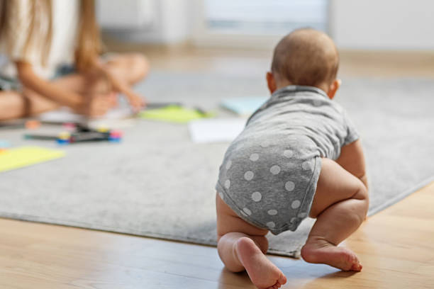 Baby wearing a gray polka dot onesie crawls on wooden floor toward carpet, with sibling blurred in background drawing on paper.