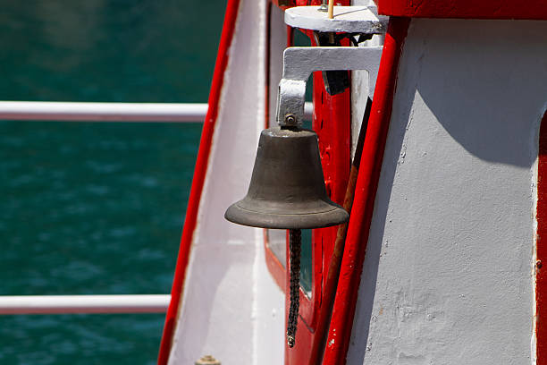 A classic bronze ship's bell is prominently featured on the side of a red and white vessel, with the vibrant turquoise sea visible in the background, hinting at a journey on the water